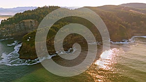 Morning light reveals cliffs, Cape Meares Lighthouse, and Oregon coast warm glow on rocks, crashing waves, and verdant forest in b