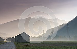 Morning mist in a mountain valley with fields and old wooden barns and lattice cross power lines and mountains in silhouette behin