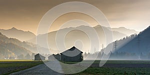 Morning mist in a mountain valley with fields and old wooden barns and lattice cross power lines and mountains in silhouette behin