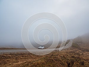 Morning mountain highway in dense fog. White car SUV with fog lights turned on is parked next to a scenic route road in a foggy