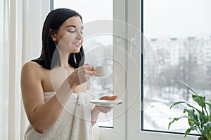 Morning portrait of young beautiful woman in bath towel with cup of fresh coffee near the window