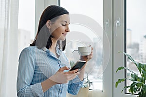 Morning portrait of young smiling woman in shirt at home near the window with cup of coffee reading text on smartphone