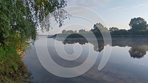 Morning on the river. A light mist and rays of sunshine. Tall grass and trees grow along the banks and are reflected in the calm w