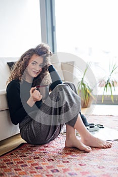 Morning routine for beautiful woman with curly hair, sitting on floor in living room, drinking cup of coffee.