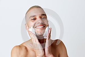 Morning routine. Portrait of smiling young man applying shaving gel, taking care after skin against white studio