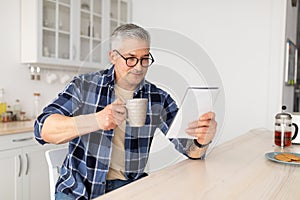 Morning routine. Senior man reading his notepad while sitting at kitchen table and drinking coffee, free space