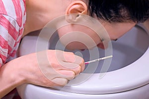 Morning sickness. Young pregnant tired woman with pregnancy test in hand is vomiting in toilet at home, closeup view.