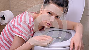 Morning sickness. Young pregnant tired woman is vomiting in toilet at home lookin at camera.