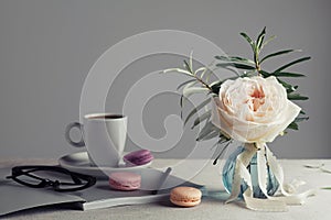 Morning still life with vintage rose in a vase, coffee and macarons on a light table. Beautiful and cozy breakfast.