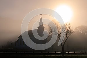 Morning sun through dense fog over small historic church and bare tree photo