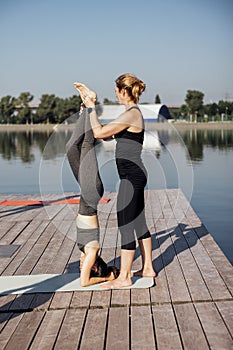 Morning yoga class on a wooden pier in the city. Young women stand in a headstand on mats. The instructor helps to do the exercise