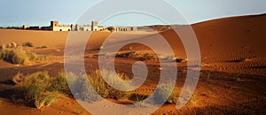 A moroccan desert scenery with sand dunes, desert grass plantation and an ancient arabic fortress on the background