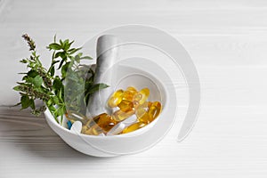 Mortar with fresh green herbs and pills on white wooden table, space for text