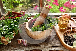 Mortar with pestle, fresh green herbs and different spices on wooden table near window