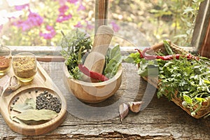 Mortar with pestle, fresh green herbs and different spices on wooden table near window