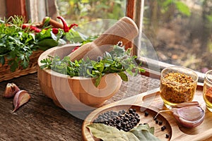 Mortar with pestle, fresh green herbs and different spices on wooden table near window