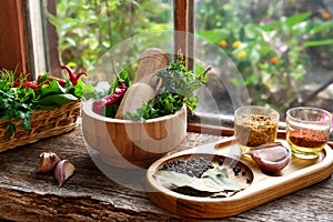 Mortar with pestle, fresh green herbs and different spices on wooden table near window