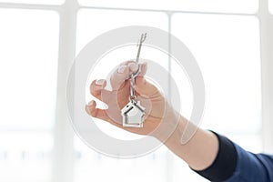 Mortgage concept. Female hand holding key with house shaped keychain. Modern light interior. Real estate, moving home or