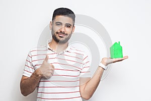 Mortgage is good. Positive attractive young man in t-shirt holding green paper small house and propose you to planning new home to