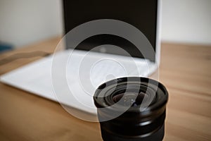 A DSLR lens on a wooden table together with a photo camera, computer