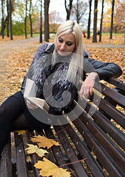 A young attractive woman wearing fur black jacket holding book sitting on the bench in falling park.