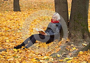 A young attractive woman wearing fur black jacket holding book sitting by the tree. Walking around autumn park.