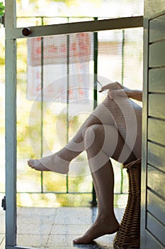 A mosquito net on the balcony door, and legs of a woman sitting in the wicker chair and smoking cigarette in a blurry background.