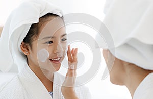 Mother adding treatment cream on the cheek to young and cute Asian girl with spa dress and head covered with a white towel. The