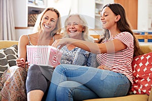 Mother With Adult Daughter And Teenage Granddaughter Eating Popcorn Watching Movie On Sofa At Home