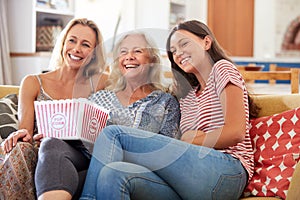 Mother With Adult Daughter And Teenage Granddaughter Eating Popcorn Watching Movie On Sofa At Home