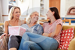 Mother With Adult Daughter And Teenage Granddaughter Eating Popcorn Watching Movie On Sofa At Home