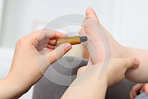 Mother applying essential oil from roller bottle onto her baby`s heel indoors, closeup