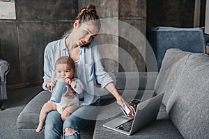 Mother with baby boy remote working and using laptop during conversation on smartphone