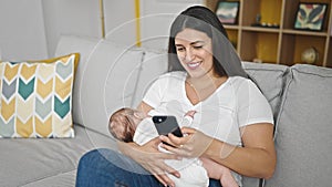 Mother and baby daughter sitting on sofa breastfeeding baby using smartphone at home