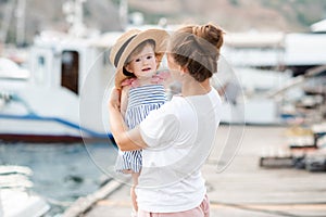 Mother with baby girl 1-2 year old with straw hat over sea and boats at background close up.