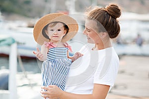 Mother with baby girl 1-2 year old with straw hat over sea and boats at background close up