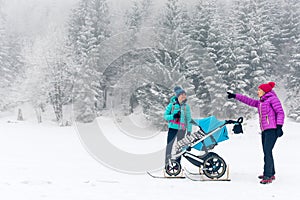 Mother with baby stroller enjoying winter forest with female friend or partner, family time. Hiking or power walking woman with