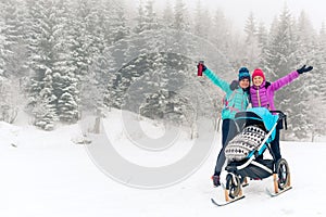 Mother with baby stroller enjoying winter forest with female friend or partner, family time. Hiking or power walking woman with