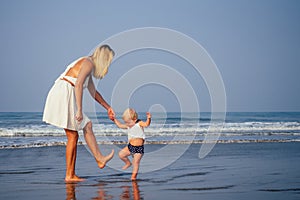Mother and baby young woman teaching her sweet toddler daughter to walk her first steps on a sandy tropical sea beach