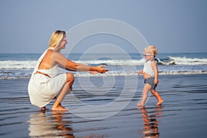 Mother and baby young woman teaching her sweet toddler daughter to walk her first steps on a sandy tropical sea beach