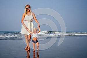 Mother and baby young woman teaching her sweet toddler daughter to walk her first steps on a sandy tropical sea beach