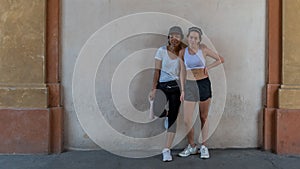 Mother with black pants and tshirt and daughter with black shorts and sports bra standing in front of wall in portico