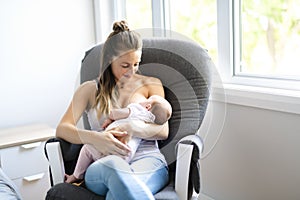 Mother is breastfeeding a baby sitting on a chair close to the window