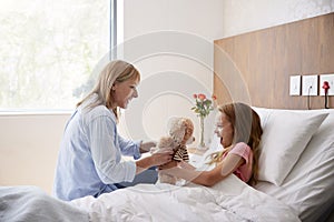 Mother Bringing Daughter Lying In Hospital Bed Gift Of Teddy Bear During Visit