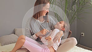 Mother with brown hair and her adorable charming daughter sitting on bed sharing joyful moments mother playfully tickles child