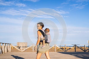 Mother carrying a baby on her back in a baby carrier next to the beach