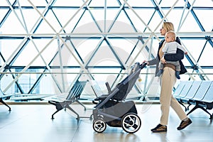 Mother carying his infant baby boy child, pushing stroller at airport departure terminal moving to boarding gates to