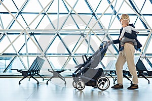 Mother carying his infant baby boy child, pushing stroller at airport departure terminal moving to boarding gates to