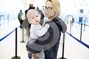 Mother carying his infant baby boy child queuing at airport terminal in passport control line at immigrations departure