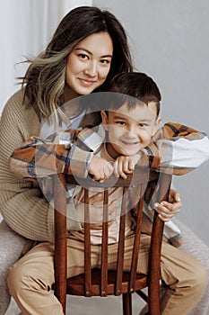 Mother and child in cozy casual clothes pose on a wooden chair. Studio shot.